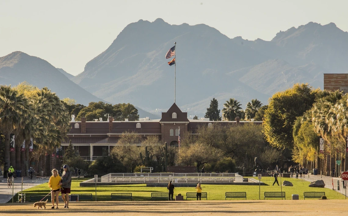 View of Old Main at the University of Arizona with the American and Arizona state flags flying atop the building. The scene is set against the backdrop of the Santa Catalina Mountains, with students walking and relaxing on the grass in the foreground, surrounded by palm trees and desert vegetation.