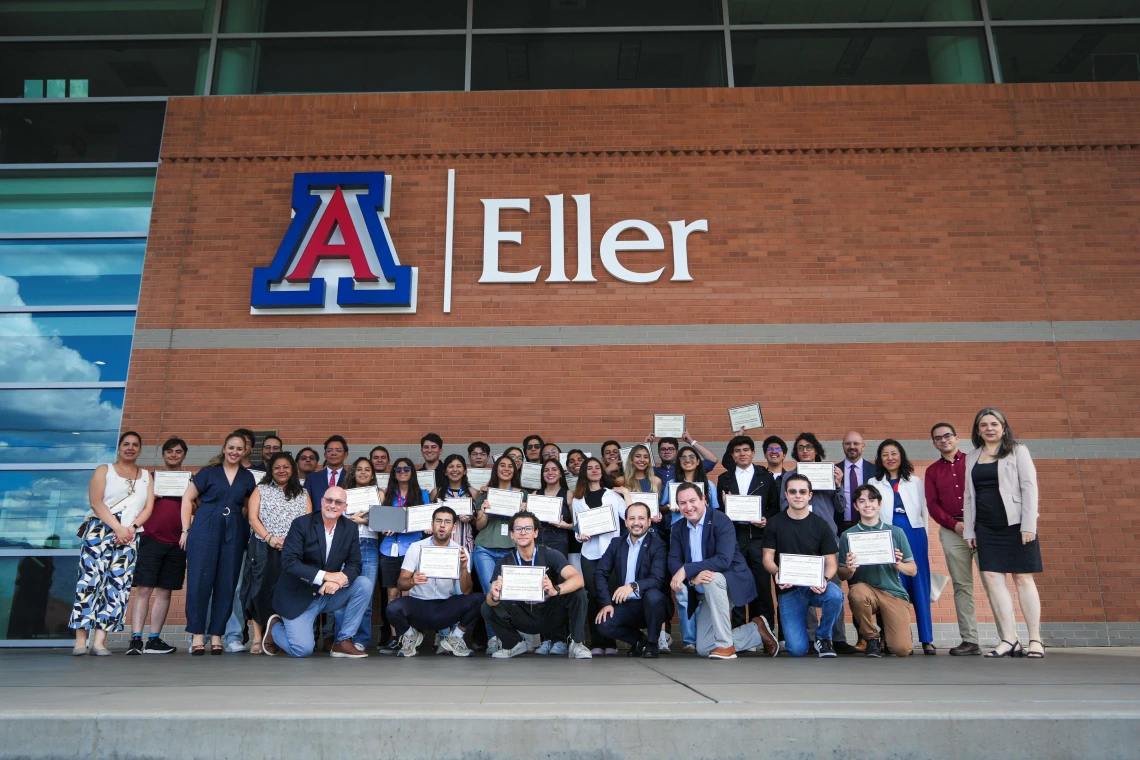 A group of students and faculty stand together in a classroom for a group photo in front of a large projector screen displaying an image of the University of Arizona campus with cactus and palm trees. Several participants wear blue lanyards, and the atmosphere appears friendly and celebratory.