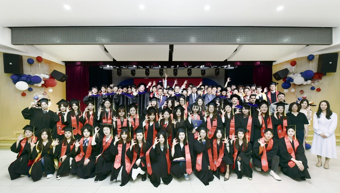A large group of graduates in black caps and gowns with red stoles pose together indoors during a commencement ceremony, making the University of Arizona "Wildcat hand sign." Faculty and administrators stand behind them on stage, with festive red, white, and blue balloons decorating the space.