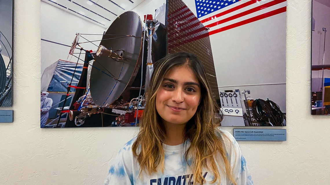 Maanyaa Kapur, wearing a light blue tie-dye “EMPATHY” T-shirt, smiles while standing indoors in front of a large photograph of the OSIRIS-REx spacecraft assembly, which features a satellite dish and an American flag.