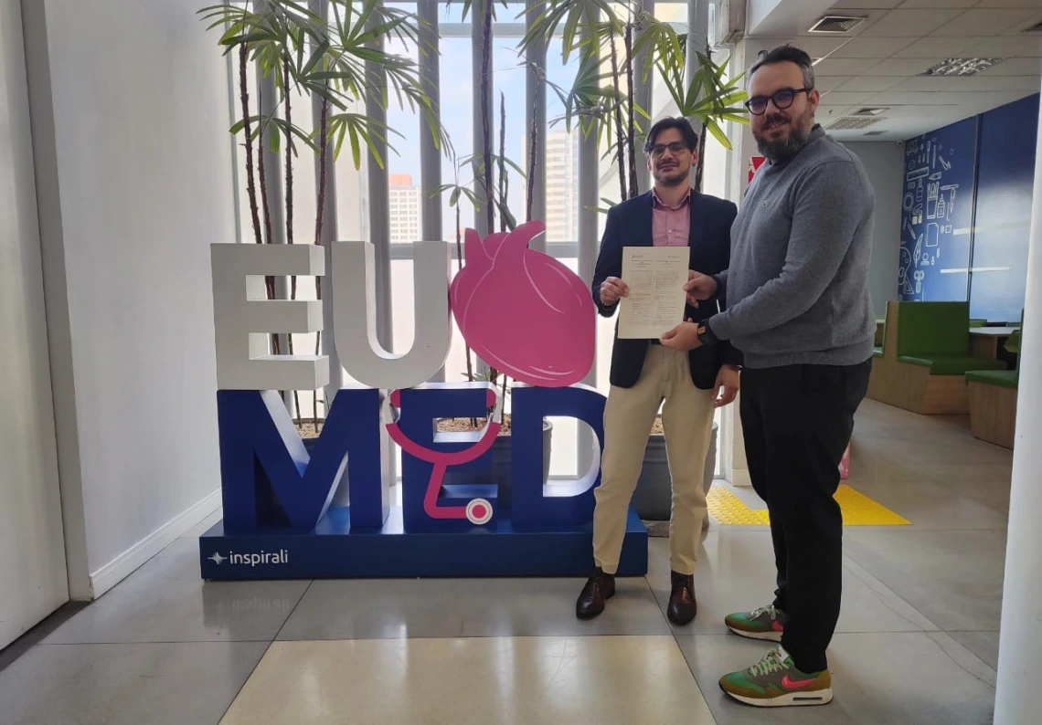 Two men stand indoors in front of a large “EU MED” sign, holding a signed document together and smiling. The sign features a pink anatomical heart and stethoscope design, with potted plants and tall windows in the background.