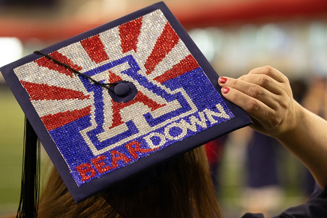 A decorated graduation cap with the University of Arizona “A” logo, red rays, and the words “BEAR DOWN” spelled out in rhinestones.