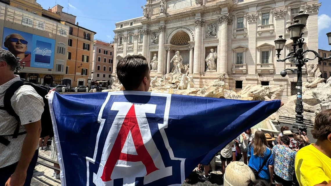 A University of Arizona student stands at the Trevi Fountain in Rome, Italy, draped in a large blue U of A flag with a red block "A" logo, surrounded by crowds of people exploring the historic landmark.