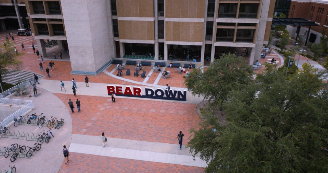 Aerial view of the University of Arizona campus plaza featuring the large red and blue “BEAR DOWN” sign, with students walking, biking, and sitting at tables outside a modern campus building.