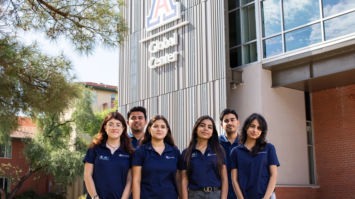 A group of seven University of Arizona Global Ambassadors stand together outside the Global Center, smiling at the camera. They are wearing matching navy polo shirts with the Arizona International logo, with the building’s large "A" logo and "Global Center" signage visible behind them.