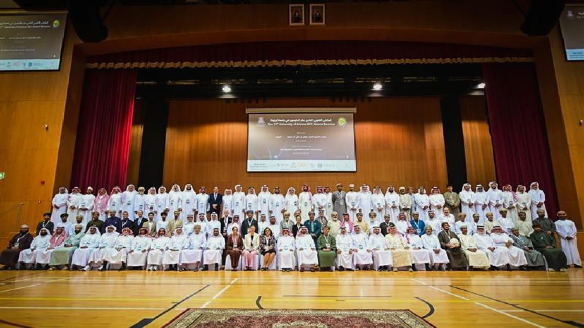 Members of the University of Arizona Gulf Cooperation Council (GCC-UA) alumni community pose for a large group photo on stage during the 11th Annual GCC Alumni Reunion in Salalah, Oman. The audience includes alumni, officials, and guests seated and standing in several rows inside an auditorium decorated with red curtains and projection screens.