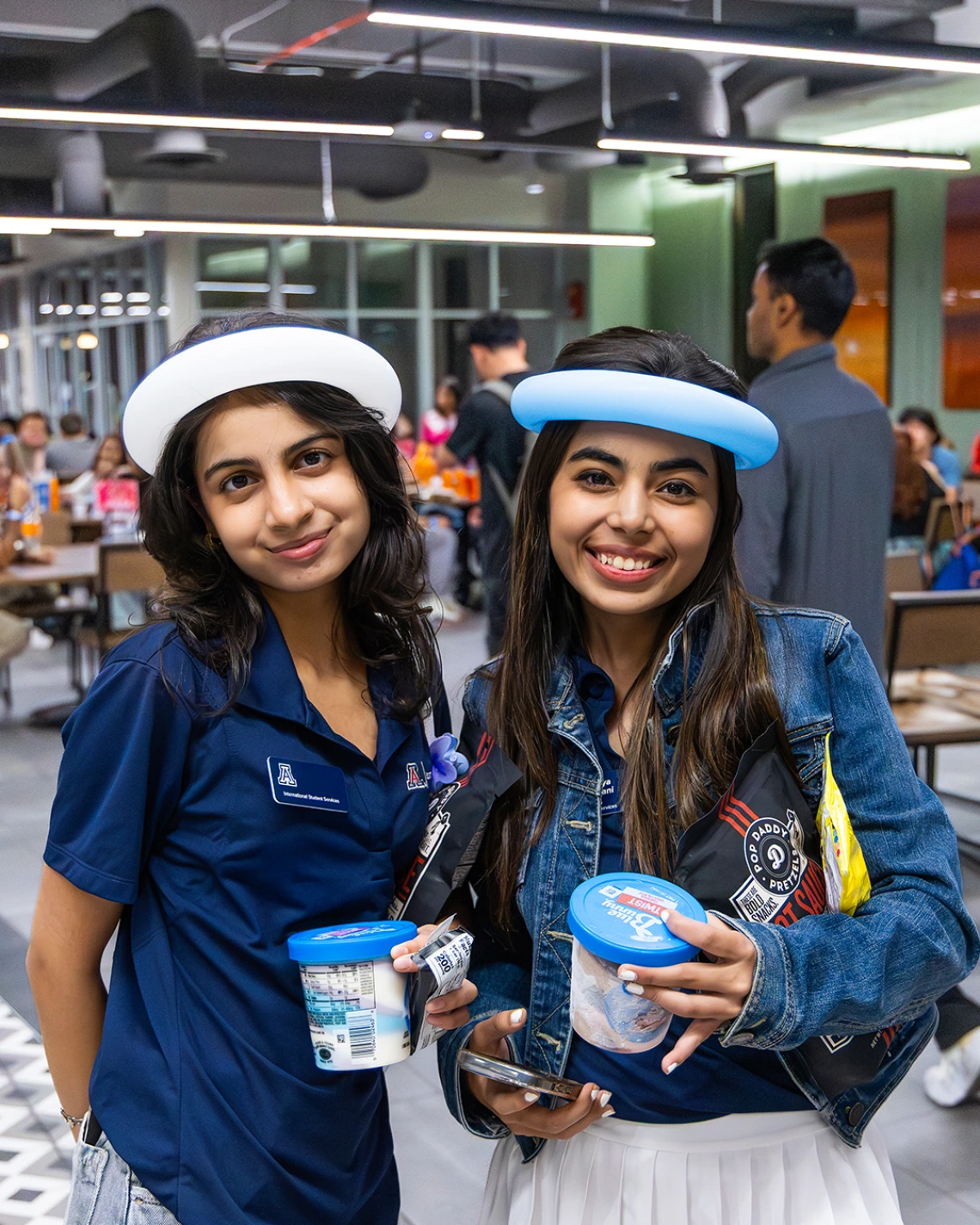 Two University of Arizona students wearing balloon hats and navy Arizona International polo shirts smile while holding ice cream and snacks at a student event inside the Global Center.