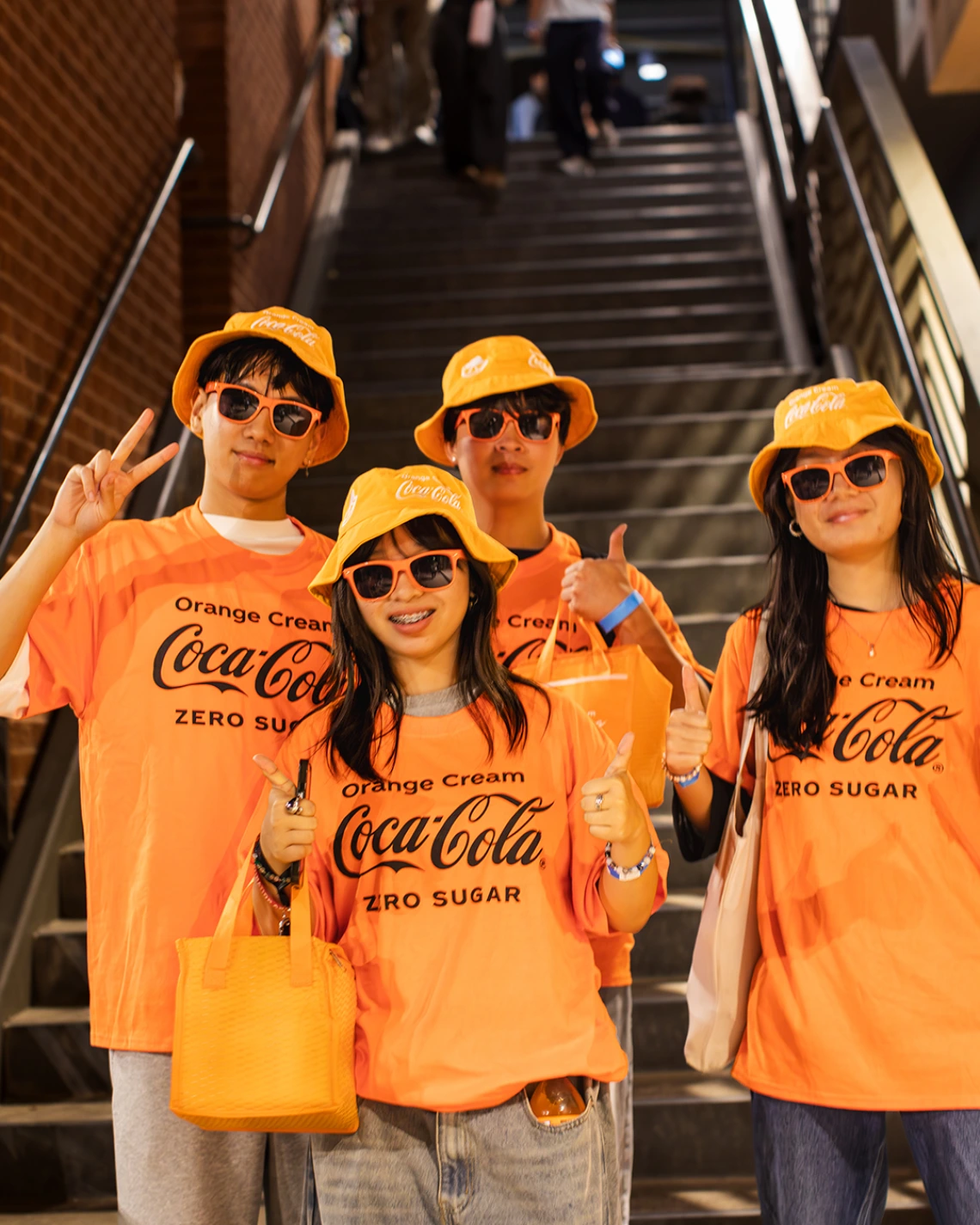 Four students wearing matching bright orange Coca-Cola “Orange Cream Zero Sugar” t-shirts, hats, and sunglasses pose together at the bottom of a staircase. They smile, give thumbs-up and peace signs, and carry orange tote bags, showing their school spirit at a campus event.