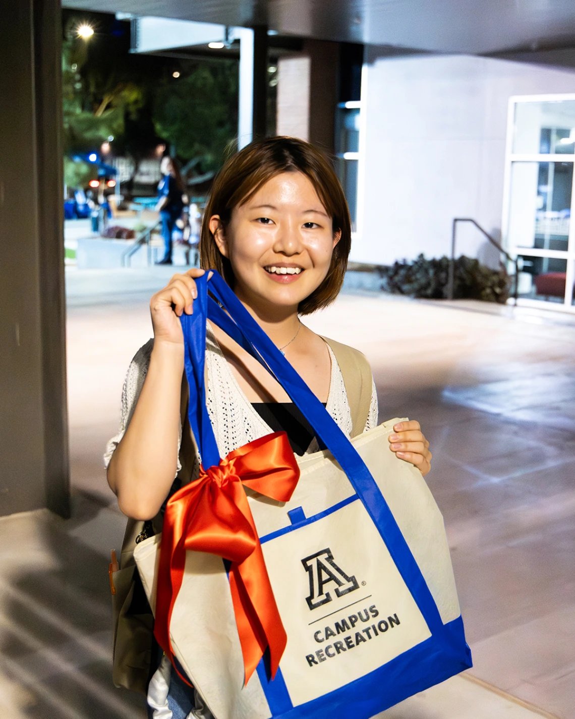 A student smiles while holding a University of Arizona Campus Recreation tote bag decorated with a large red bow during an evening event.
