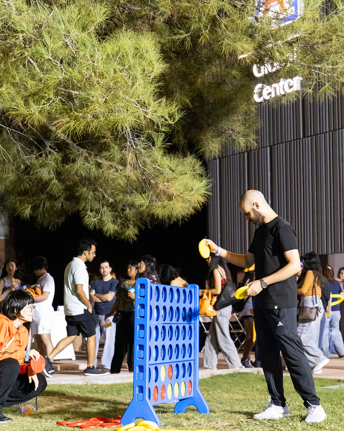Students play a giant Connect Four game on the lawn outside the University of Arizona Global Center during a nighttime event, with other attendees mingling in the background.