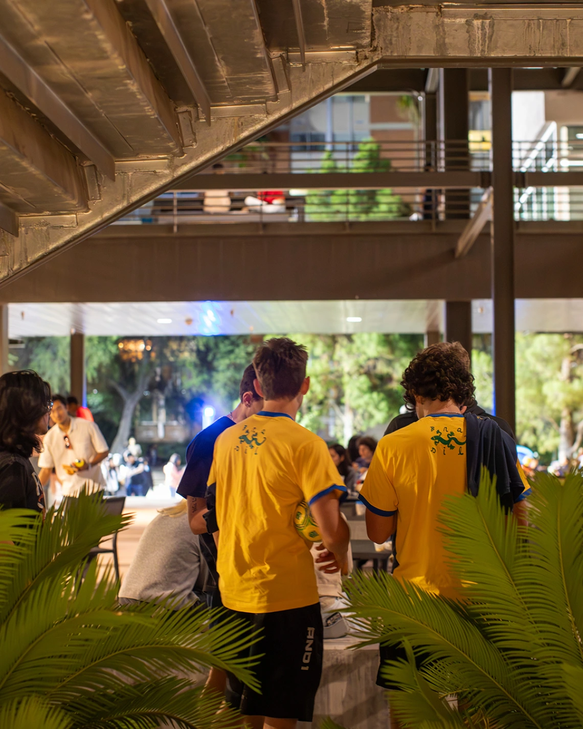 A group of students wearing matching bright yellow t-shirts socialize under an outdoor structure during a nighttime event at the University of Arizona Global Center. Green plants frame the foreground while other students gather and chat in the background.