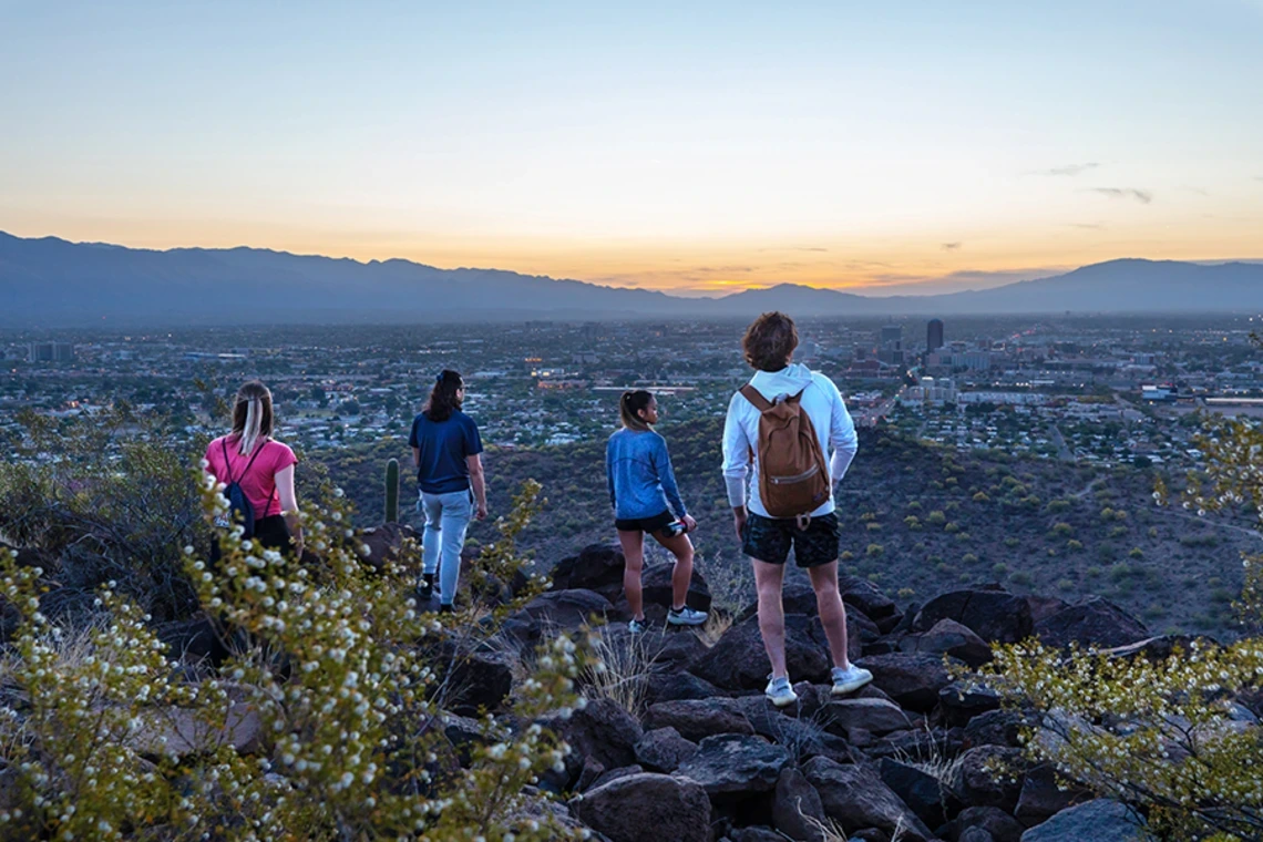 Four people stand on rocky terrain at sunset, overlooking the city of Tucson, Arizona. The view includes the sprawling cityscape below with distant mountains and a colorful sky.