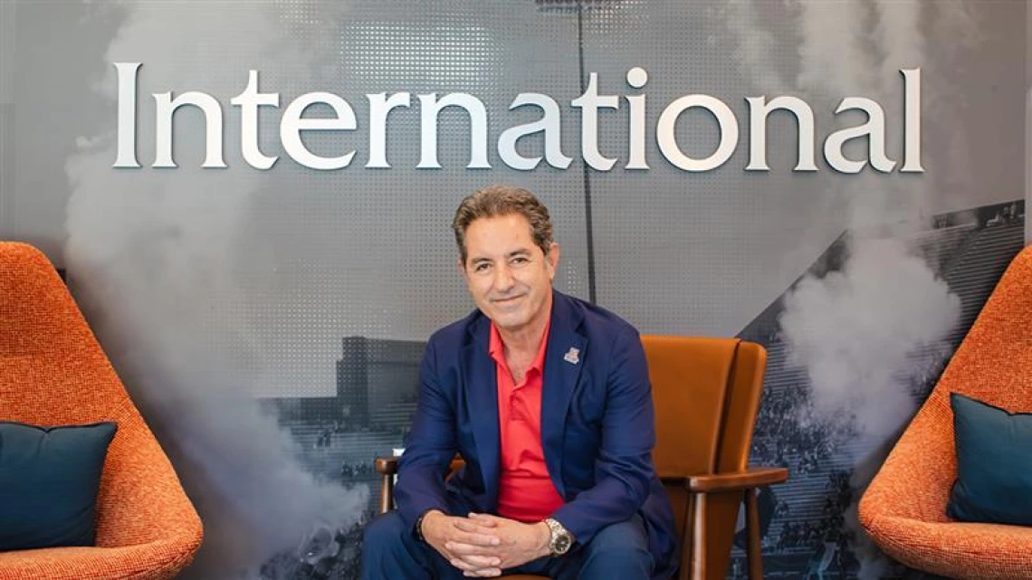 Hassan Hijazi, Senior Assistant Vice President for University of Arizona International, sits in a chair in the Arizona International office. He is wearing a navy blue suit with a red shirt, smiling at the camera. Behind him, the word “International” is displayed prominently on the wall.