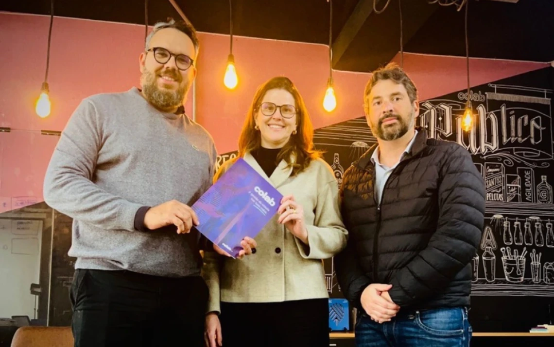 Three people stand together indoors under warm hanging lights, smiling at the camera. The person on the left holds a purple and blue booklet. The background features a chalkboard-style mural with text and illustrations, giving the space a casual, creative atmosphere.