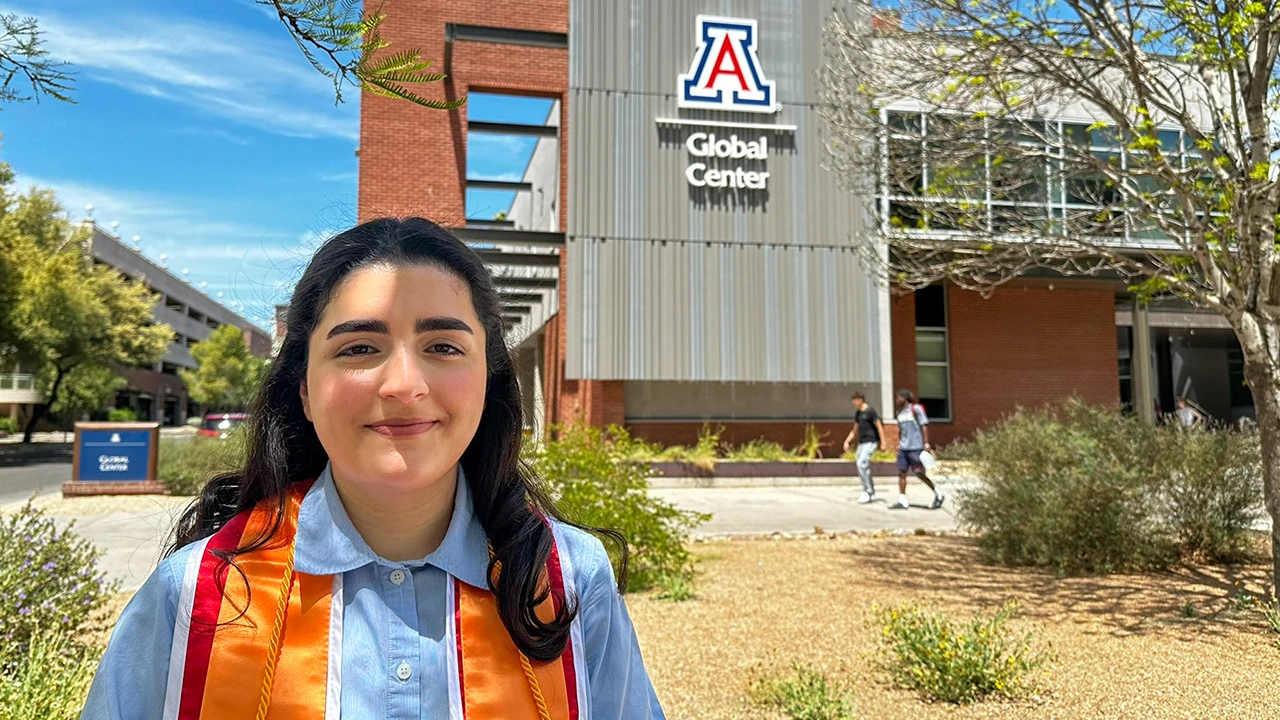 A young woman wearing a light blue shirt and an orange and red graduation stole smiles in front of the University of Arizona Global Center. The building behind her prominently features the U of A logo and the words “Global Center” on a modern metal and brick facade. Trees, desert landscaping, and a few students walking in the background reflect a sunny day on campus, highlighting a celebratory graduation moment.