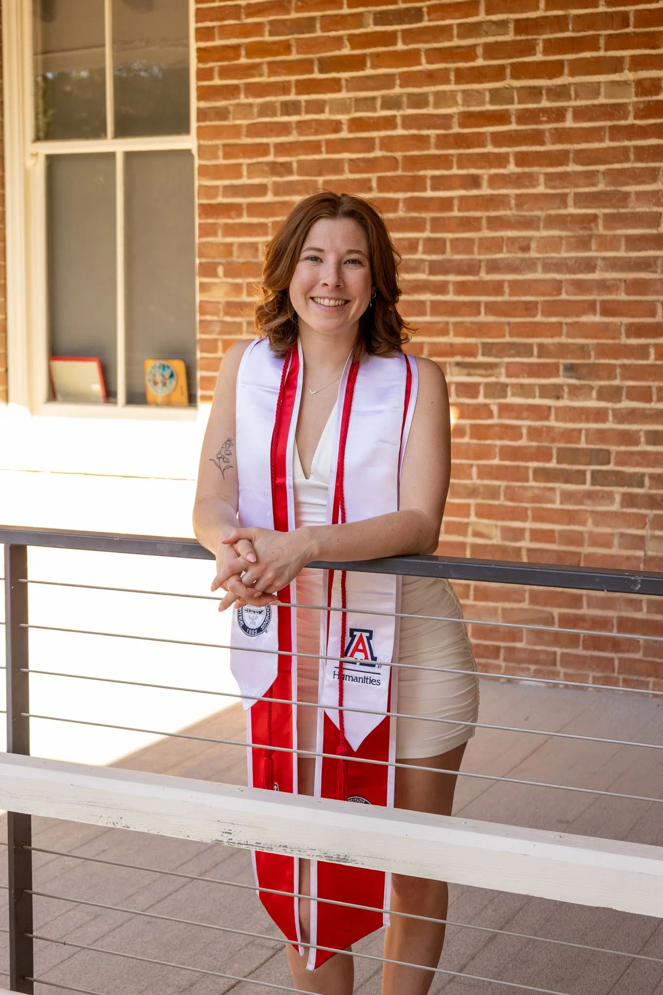 A young woman wearing a white dress and red-and-white graduation stole stands smiling and leaning on a railing in front of a red brick wall. Her stoles feature the University of Arizona and College of Humanities logos, as well as honor cords. Sunlight filters in from the left, casting a warm glow over the scene and highlighting her achievement and cheerful expression. A tattoo is visible on her upper arm, and framed mementos can be seen on the windowsill behind her.