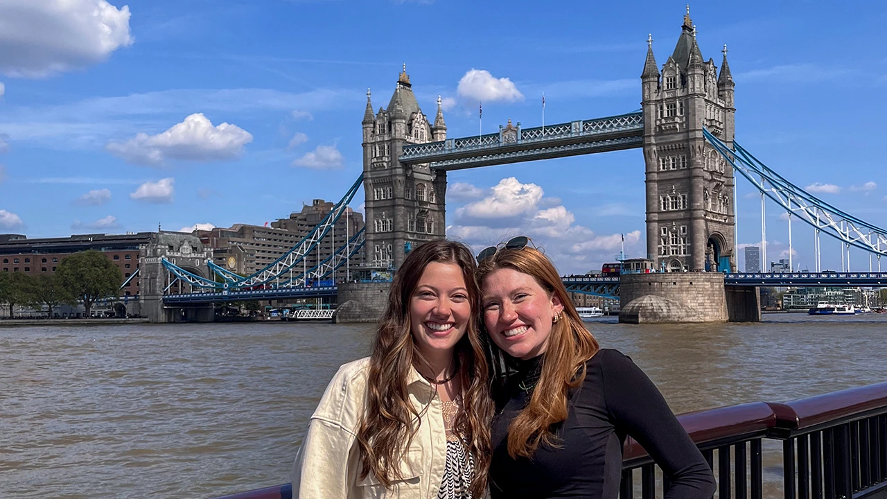 Two women smiling and standing close together in front of London’s Tower Bridge on a sunny day, with the River Thames and the iconic twin towers of the bridge in the background.