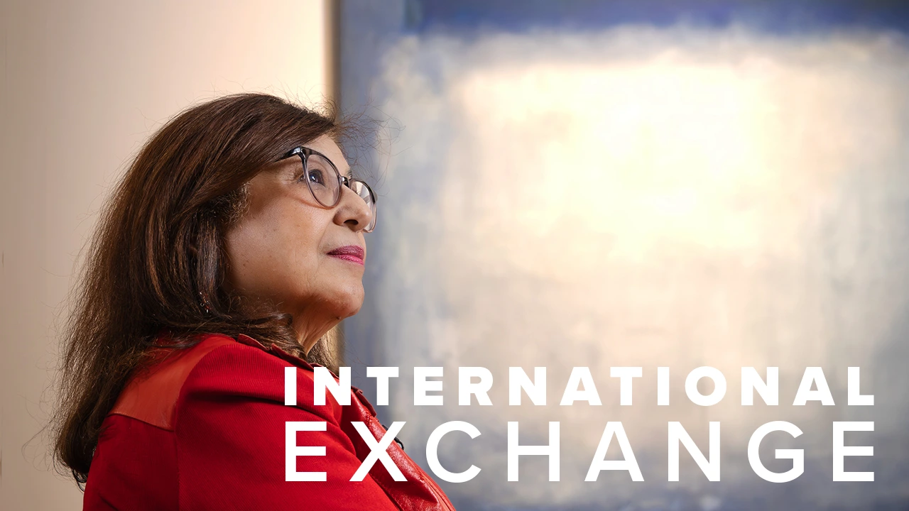 Dr. Iman Hakim, dean of the University of Arizona Mel and Enid Zuckerman College of Public Health, looks thoughtful while gazing upward. She is wearing glasses and a red outfit, with the words "International Exchange" displayed in bold white text beside her.