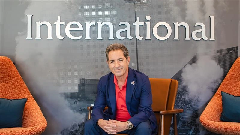 Hassan Hijazi, Senior Assistant Vice President for University of Arizona International, sits in a chair in the Arizona International office. He is wearing a navy blue suit with a red shirt, smiling at the camera. Behind him, the word “International” is displayed prominently on the wall.