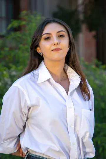 Photo of Montessa White, Visual Designer for Arizona International. They have brown hair and a white button up shirt. The background is blurred trees, shrubs and a red brick building.