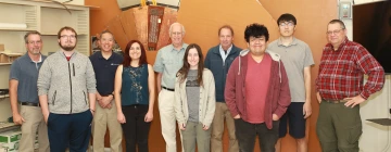 A group of eleven individuals—comprising a mix of students, faculty, and researchers—stand side by side smiling for a photo in a lab or workspace. Behind them is a large, intricate copper-colored component, likely a particle detector or scientific instrument. The group appears to be part of a research team, possibly involved with the ATLAS experiment or related particle physics project. They are dressed in casual and business-casual attire, and the environment reflects an academic or engineering setting.