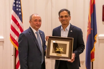 Two men in formal attire stand side by side smiling, with one of them holding a framed artwork featuring two golden ibexes. The man on the right wears a University of Arizona lapel pin on his navy blazer. Behind them, the U.S. flag and the Arizona state flag are visible, indicating a formal or diplomatic event. The setting appears to be an indoor ceremony or meeting with cultural or institutional significance.