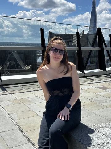A young woman wearing a strapless black outfit and sunglasses sits on a rooftop terrace with a view of London’s skyline behind her. The iconic Shard building rises prominently in the background under a partly cloudy sky. She smiles gently at the camera, enjoying the sunny weather and urban scenery. Glass railings and modern architecture frame the elevated setting.