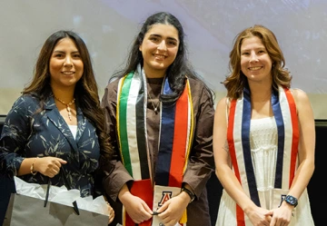 Three young women stand side by side smiling at the U of A Global Graduation celebration. Two of them wear stoles representing the University of Arizona and their international or study abroad affiliations—one with a multicolored stole including the Mexican and Indian flags, and the other with a red, white, and blue stole. The woman on the left wears a navy floral blazer and holds a gift bag. The setting is festive and academic, highlighting recognition and celebration of their achievements.