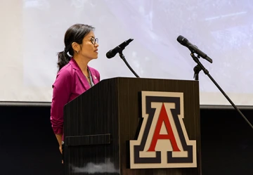 A woman in a magenta blazer stands at a podium adorned with the U of A logo, speaking into two microphones. She faces an audience, with a projection screen visible behind her displaying a faint image. The setting appears to be a formal event, likely the U of A Global Graduation ceremony, where she is delivering remarks or a welcome address. Her expression is composed and engaged.