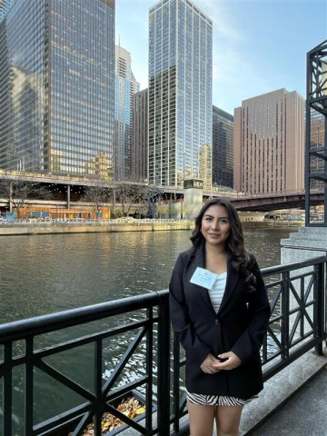 A young woman in a black blazer and patterned skirt stands smiling by a riverside railing in an urban setting. She wears a name badge and poses confidently with tall glass and steel skyscrapers and a bridge in the background. The river reflects the buildings and soft evening light, suggesting a professional event or conference in a city like Chicago. The scene captures a blend of personal achievement and metropolitan energy.