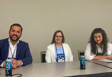 Three international education professionals sit at a panel table during a NAFSA conference session. Each has a can of water in front of them, and they are smiling warmly. The panelists are dressed in business attire and appear ready to speak or answer questions in a professional, conference setting.