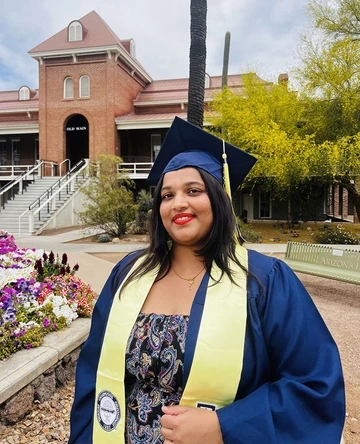 Ashwini R. Jannu, MSIS ’25, poses in cap and gown in front of Old Main at the University of Arizona. She wears a gold stole and a floral dress, smiling beside colorful spring flowers.