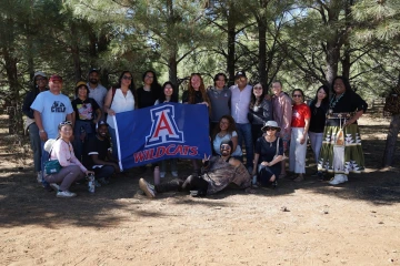 A diverse group of people pose together in a shaded forest clearing, holding a University of Arizona "WILDCATS" flag with the red, white, and blue "A" logo. The group includes students, staff, and community members, some in casual outdoor attire and one woman in traditional Native American dress. Everyone is smiling, with one man playfully posing in front of the group on the ground, flashing a peace sign. Sunlight filters through the pine trees behind them.
