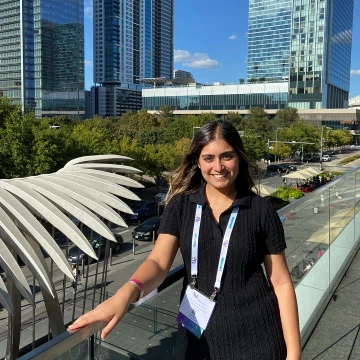 Maanyaa Kapur stands on an outdoor balcony with modern high-rise buildings and green trees in the background. She is wearing a black short-sleeved dress and a conference badge lanyard, smiling at the camera under a sunny blue sky.