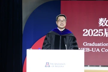A speaker in academic regalia stands at a podium with the University of Arizona logo, delivering remarks at the 2025 graduation ceremony for the CUEB-UA cohort.