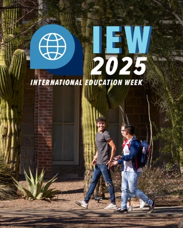 Two University of Arizona students walk past tall saguaro cacti on campus during a sunny day. The image includes the International Education Week 2025 logo and text.