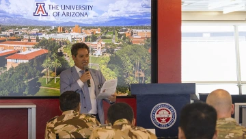 Hassan speaks into a microphone while holding a sheet of paper at a podium in front of an audience wearing camouflage uniforms. Behind him is a large screen displaying an aerial view of the University of Arizona campus and its logo.