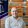 An older man with gray hair and a friendly smile sits in front of a modern backdrop of horizontal orange slats. He wears a light checkered button-down shirt and is positioned beside a banner displaying scales of justice and the words “College of Law,” suggesting a connection to a law school. The setting appears to be an academic or institutional space.