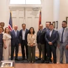 A group of fourteen individuals, including men and women, pose together in a formal setting in front of a pair of white double doors flanked by the U.S. and Arizona state flags. Most individuals are dressed in business attire, suggesting a diplomatic or professional delegation. In the foreground, a framed artwork rests on a wooden table, indicating a ceremonial exchange or gift presentation. The room is well-lit and adorned with framed certificates and plaques, reinforcing the official and celebratory natur