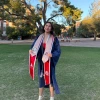 A young woman wearing a navy blue graduation gown and several stoles stands on a grassy area of the University of Arizona campus during golden hour. She smiles confidently, holding the ends of her stoles, which feature the University of Arizona, Eller College of Management, and other honors logos. She wears a red dress and white high-heeled sandals. Behind her, large trees and red-brick campus buildings are bathed in warm sunlight, creating a celebratory and serene atmosphere.