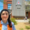 A young woman wearing a light blue shirt and an orange and red graduation stole smiles in front of the University of Arizona Global Center. The building behind her prominently features the U of A logo and the words “Global Center” on a modern metal and brick facade. Trees, desert landscaping, and a few students walking in the background reflect a sunny day on campus, highlighting a celebratory graduation moment.