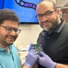Two researchers smile while holding a small green circuit board labeled “Graphene” in a laboratory setting. Both are wearing glasses and blue gloves, with one in a teal polo shirt and the other in a black collared shirt. A monitor in the background displays scientific imagery and the words “Hassan group.” The setting suggests an academic research lab focused on advanced electronics or materials science.