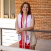 A young woman wearing a white dress and red-and-white graduation stole stands smiling and leaning on a railing in front of a red brick wall. Her stoles feature the University of Arizona and College of Humanities logos, as well as honor cords. Sunlight filters in from the left, casting a warm glow over the scene and highlighting her achievement and cheerful expression. A tattoo is visible on her upper arm, and framed mementos can be seen on the windowsill behind her.