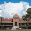 A front-facing view of Old Main, the historic red-brick building at the University of Arizona, framed by palm trees and desert greenery. A wide staircase leads to the main entrance under a prominent gabled roof. Puffy white clouds and a bright blue sky form a dramatic backdrop, highlighting the architectural charm and heritage of the university’s oldest building.