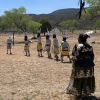 A group of Indigenous dancers dressed in traditional attire perform an outdoor ceremonial dance on dry grassland surrounded by trees and mesas. Male dancers wear body paint, headpieces, and yellow garments, while female dancers in colorful skirts and sashes observe or participate. The scene is set in a rural landscape under a clear blue sky.