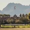 View of Old Main at the University of Arizona with the American and Arizona state flags flying atop the building. The scene is set against the backdrop of the Santa Catalina Mountains, with students walking and relaxing on the grass in the foreground, surrounded by palm trees and desert vegetation.