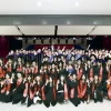 A large group of graduates in black caps and gowns with red stoles pose together indoors during a commencement ceremony, making the University of Arizona "Wildcat hand sign." Faculty and administrators stand behind them on stage, with festive red, white, and blue balloons decorating the space.