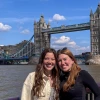 Two women smiling and standing close together in front of London’s Tower Bridge on a sunny day, with the River Thames and the iconic twin towers of the bridge in the background.