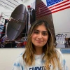 Maanyaa Kapur, wearing a light blue tie-dye “EMPATHY” T-shirt, smiles while standing indoors in front of a large photograph of the OSIRIS-REx spacecraft assembly, which features a satellite dish and an American flag.