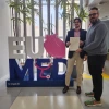 Two men stand indoors in front of a large “EU MED” sign, holding a signed document together and smiling. The sign features a pink anatomical heart and stethoscope design, with potted plants and tall windows in the background.